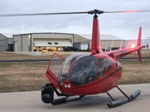 Red helicopter with a camera gimbal on the skid, rotors spinning in front of hangars