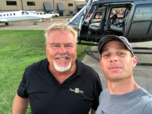 Two men on an airfield in front of a dark helicopter with a business jet behind them