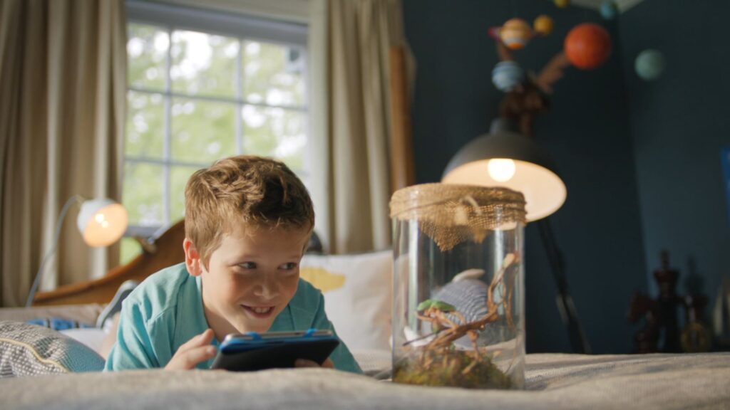 Boy on a bed looking at a tablet beside a lizard in a jar