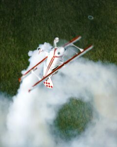 White and red biplane trailing smoke over green fields
