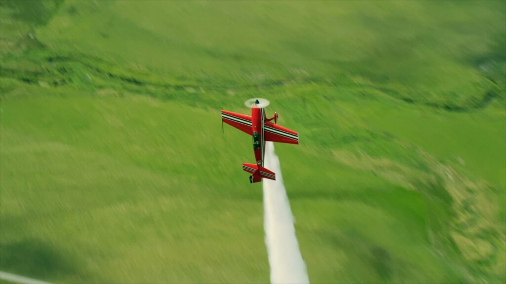 Red and silver aerobatic biplane trailing smoke over farmland