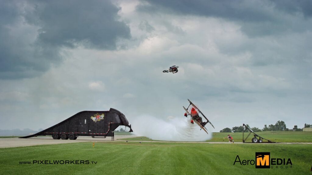 Motorcycle jumping a ramp as an aerobatic biplane passes low at an airshow