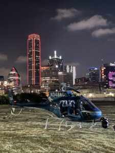 Blue helicopter with a camera gimbal parked at night with the city skyline behind it