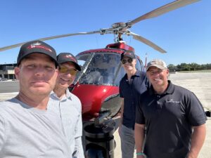 Four men standing in front of a maroon helicopter with a nose gimbal