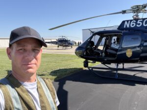 Man in a flight vest on the tarmac in front of a dark helicopter with a nose gimbal